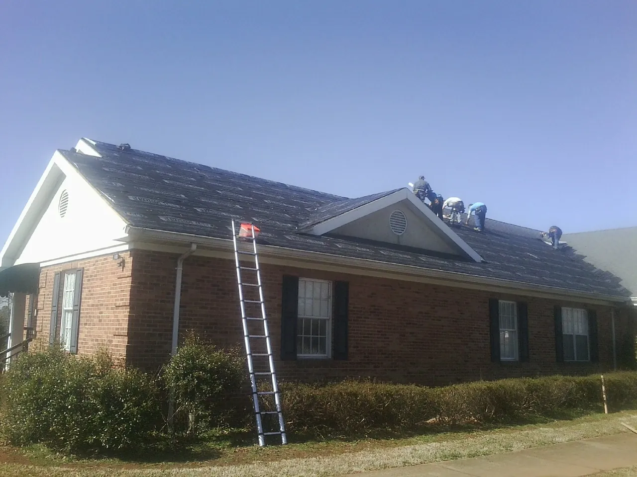 Crew tearing shingles off a brick ranch roof with a ladder set — Metal Roof Repair work by our crew in Jud