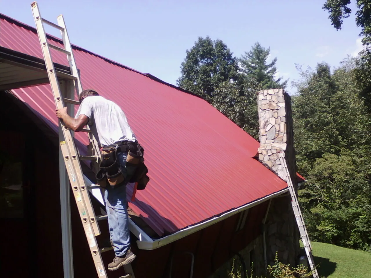 Roofer on a ladder beside a red metal roof and stone chimney — Soffit & Fascia Repair by Roofing Brothers in Jud